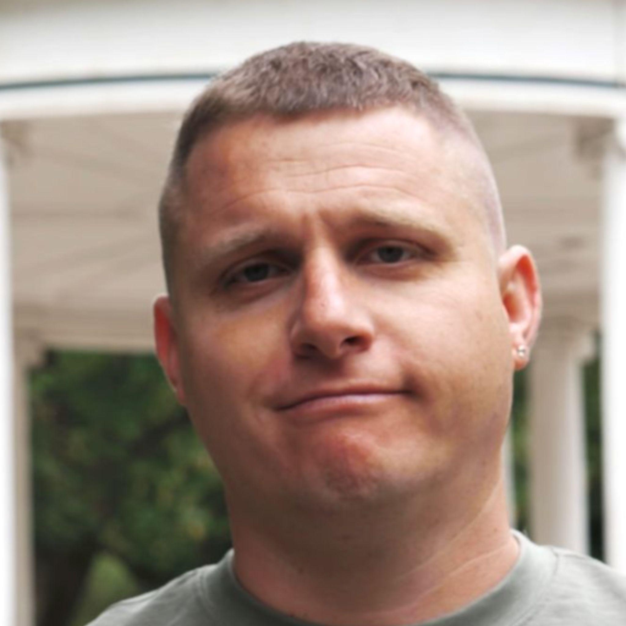 A man with a neutral expression stands in front of a white bandstand with columns, set against a backdrop of green trees.