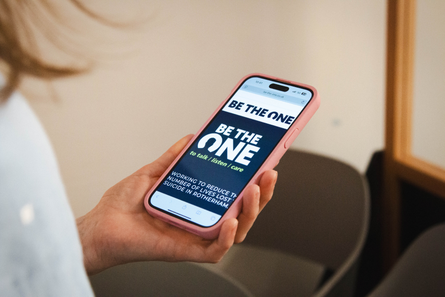 A person holds a smartphone displaying the text "Be the One to talk, listen, care." The phone has a pink case and is viewed in a room with chairs. The tone is encouraging and supportive.