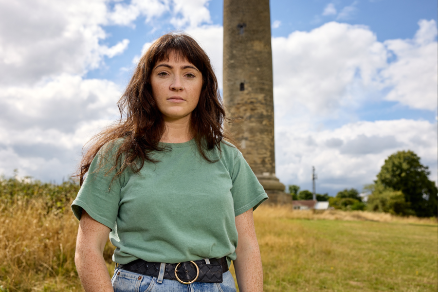 A woman with brown hair, wearing a green shirt and jeans, stands confidently in a grassy field with a tall stone tower in the background under a partly cloudy sky.