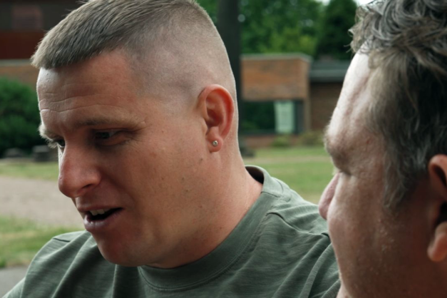 Two men outdoors, talking intently amidst greenery; a brick building is blurred in the background.