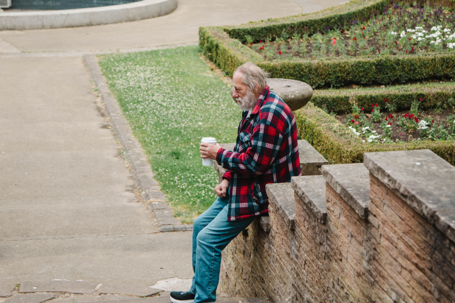 An elderly man in a red plaid jacket sits on stone steps in a garden, holding a coffee cup. The mood is peaceful, with neatly trimmed hedges and scattered flowers.