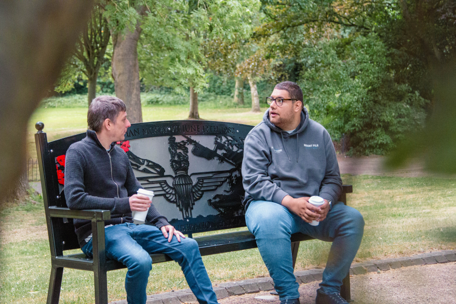 Two men sit on a decorative park bench, holding coffee cups, engaged in conversation. They are surrounded by lush greenery, creating a relaxed atmosphere.