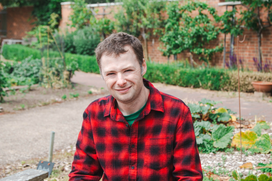 A person in a red flannel shirt smiles in a garden with neatly trimmed hedges and plants. The scene is bright and lively, conveying a cheerful mood.